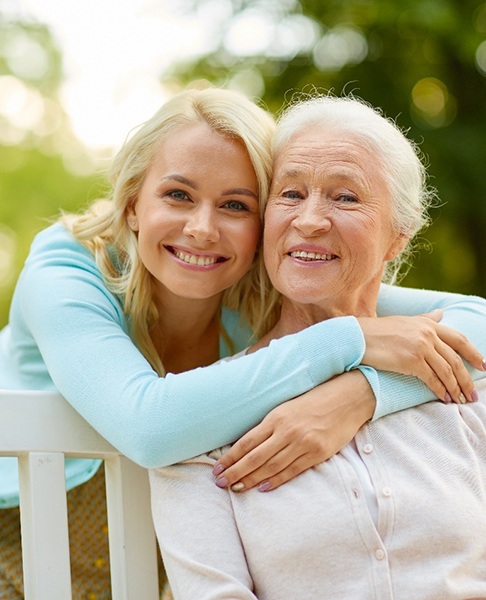 An older woman with dental implants in Dallas is sitting on a bench while another woman hugs her and smiles