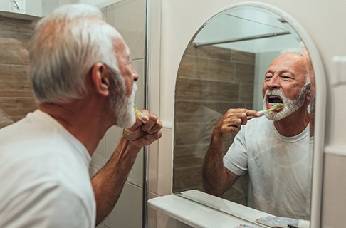 An older man is standing in front of his mirror brushing his teeth in his bathroom in Dallas
