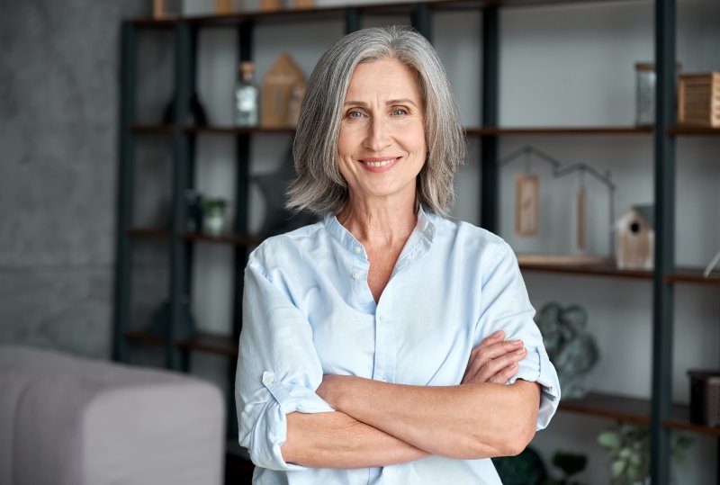 A middle-aged woman standing with her arms crossed and smiling after receiving dental implants